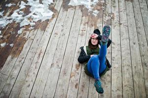 Young girl wear on long green sweatshirt, jeans and black headwear sitting on wooden floor with snow. photo