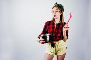 Young funny housewife in checkered shirt and yellow shorts pin up style with saucepan and kitchen spoon isolated on white background. photo