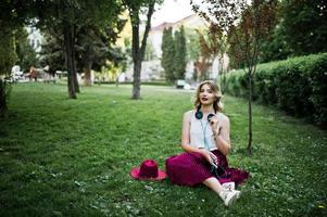 Fashionable and beautiful blonde model girl in stylish red velvet velour skirt, white blouse and hat, sitting on green grass at park with phone and earphones. photo