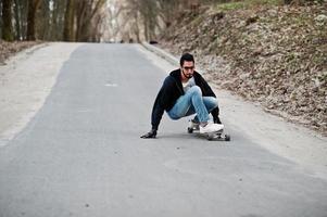 hombre árabe de estilo callejero en anteojos con longboard longboarding por el camino. foto