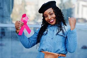 The power to fight. Stylish fashionable african american women in jeans wear and black beret against modern building with pink ribbon for breast cancer. photo