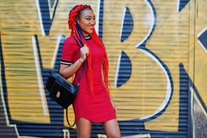 linda y delgada chica afroamericana vestida de rojo con rastas y mochila posada al aire libre en la pared de graffiti de fondo de la calle. elegante modelo negro. foto