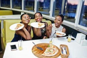 tres mujeres africanas vestidas posando en el restaurante, comiendo pizza y bebiendo jugo. foto