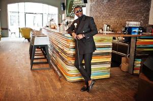 elegante caballero afroamericano con elegante chaqueta negra y gafas de sol, sosteniendo un bastón retro como matraz de caña o bastón basculante con mango de bola de diamante dorado. rico hombre afro de moda. foto