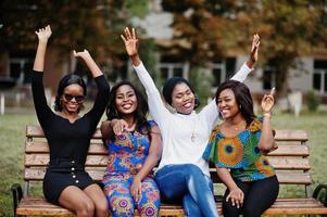 grupo de cuatro chicas afroamericanas sentadas en un banco al aire libre y poniendo las manos en el aire. foto