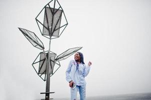 Stylish african american girl with dreads holding mobile phone at hand, outdoor against solar batteries at snowy weather. photo