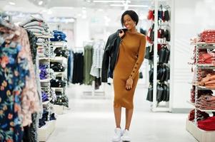 Thin african american woman in brown tunic dress and black leather jacket posed at clothes store. It's time for shopping. photo