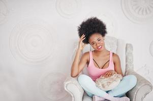 joven mujer afroamericana en camiseta rosa sentada en una silla contra la pared blanca. foto