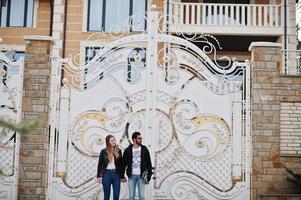 Cool multiracial couple standing with longboard against tall gates. photo