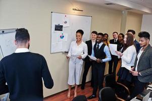 Mixed race business coach presenting report standing near whiteboard pointing on sales statistic shown on diagram and chart teach diverse company members gathered together in conference room. photo