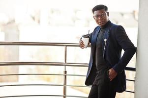 hombre afroamericano de moda con traje y gafas con una taza de café en las manos posado en el balcón de la oficina. foto