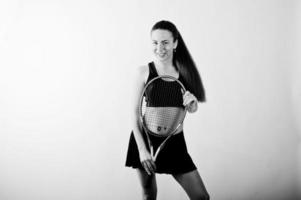 Black and white portrait of beautiful young woman player in sports clothes holding tennis racket while standing against white background. photo