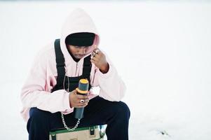 African american fisherman with thermos sitting on frozen lake. Winter fishing. photo