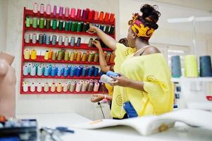 Two african dressmaker woman sews clothes on sewing machine and selects threads at tailor office. Black seamstress girls. photo