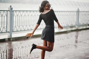 African american girl in sunglasses, black clothes and shirt posed outdoor. Fashionable black woman against rain of fountains. photo