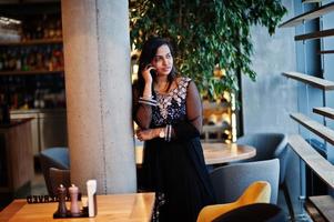 Pretty indian girl in black saree dress posed at restaurant with mobile phone at hand. photo