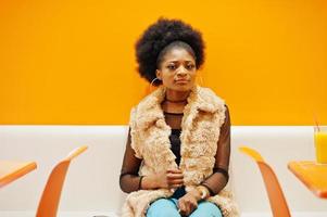 African woman with afro hair sitting on cafe against orange wall. photo