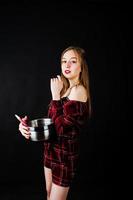 Young housewife in checkered dress with saucepan and kitchen spoon isolated on black background. photo