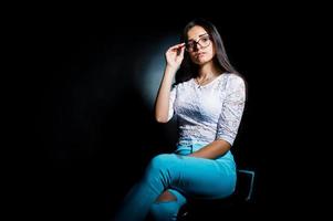 Portrait of an attractive young woman in white top and blue pants sitting posing with her glasses in the dark. photo