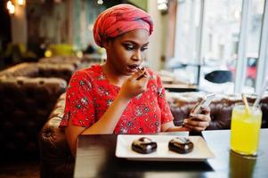 Stylish african woman in red shirt and hat posed indoor cafe, eat chocolate dessert cakes and looking at mobile phone. photo