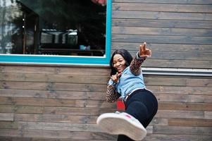 Hipster african american girl wearing jeans shirt with leopard sleeves simulates a kick at street against wooden house with windows. photo