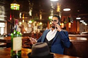 un apuesto hombre árabe bien vestido con un vaso de whisky y un cigarro sostiene un teléfono móvil, posado en el pub. foto
