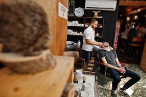 Young bearded man washing head by hairdresser while sitting in chair at barbershop. Barber soul. photo