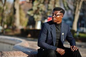 un hombre afroamericano de aspecto asombroso usa un blazer azul con broche, cuello alto negro y anteojos posados en la calle. chico negro de moda. foto