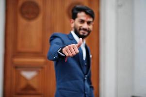 hombre indio de barba elegante con bindi en la frente, vestido con traje azul posado al aire libre contra la puerta del edificio y mostrando el pulgar hacia arriba. foto