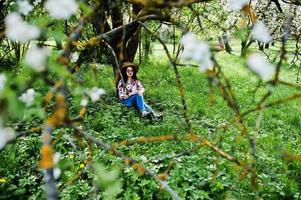 Spring portrait of brunette girl in pink glasses and hat at green blossom garden. photo