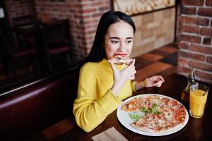 Funny brunette girl in yellow sweater eating pizza at restaurant. photo