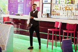 Young handsome african man wearing white shirt, black vest and bow tie with wallet purse posed against bar counter at night club. photo