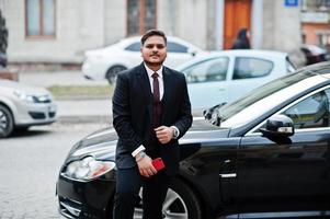 Stylish indian businessman in formal wear with mobile phone standing against black business car on street of city. photo