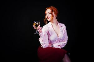 Gorgeous red haired girl in pink blouse and red skirt with glass of wine at hand isolated on black. photo