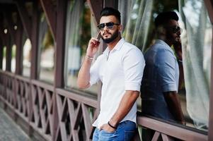 Stylish tall arabian man model in white shirt, jeans and sunglasses posed at street of city. Beard attractive arab guy speaking on mobile phone. photo