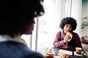 Two curly hair african american woman wear on sweaters sits at table cafe , eat croissant and drink tea. photo