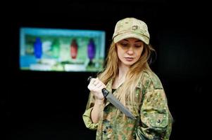 Military girl in camouflage uniform with knife at hand against army background on shooting range.