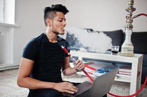 hombre indio guapo y de moda en negro sentado en la habitación, fumando narguile y trabajando en la computadora portátil. foto