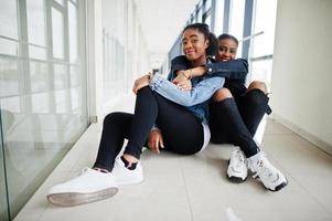 Two african woman friends in jeans jacket sitting indoor together. photo