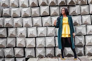 Stylish african american woman at green coat and yellow dress posed against stone texture background. photo