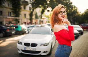 Attractive redhaired woman in eyeglasses, wear on red blouse and jeans skirt posing at street against white sport car. photo