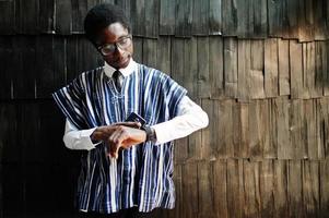 African businessman in traditional clothes and glasses with mobile phone at hand looking at his watches against wooden wall. photo