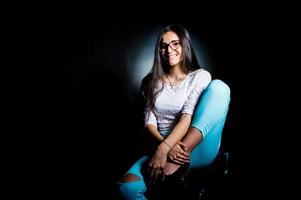 Portrait of an attractive young woman in white top and blue pants sitting posing with her glasses in the dark. photo