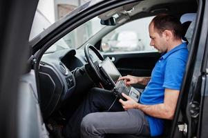 Car repair and maintenance theme. Electric mechanic in uniform working in auto service, making car diagnostics using obd device with laptop. photo