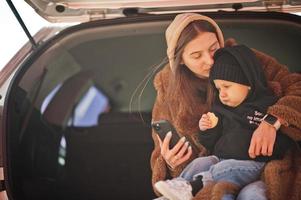 Young mother and child sitting in the trunk of a car and looking at mobile phone. Safety driving concept. photo