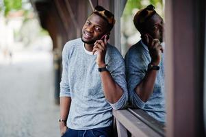 Stylish african american boy on gray sweater and black sunglasses posed on street and speaking on phone. Fashionable black guy. photo