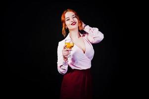 Gorgeous red haired girl in pink blouse and red skirt with glass of wine at hand isolated on black. photo