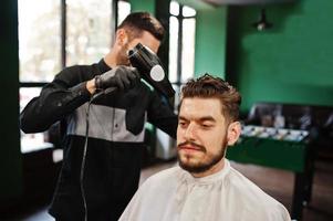 Handsome bearded man at the barbershop, barber at work, using hair dryer. photo