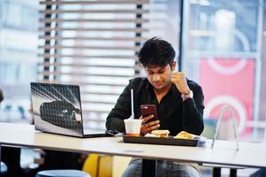 Stylish indian man sitting at fast food cafe against his laptop with mobile phone at hand. photo