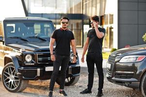 Two asian brothers man wear on all black posed near suv cars. photo